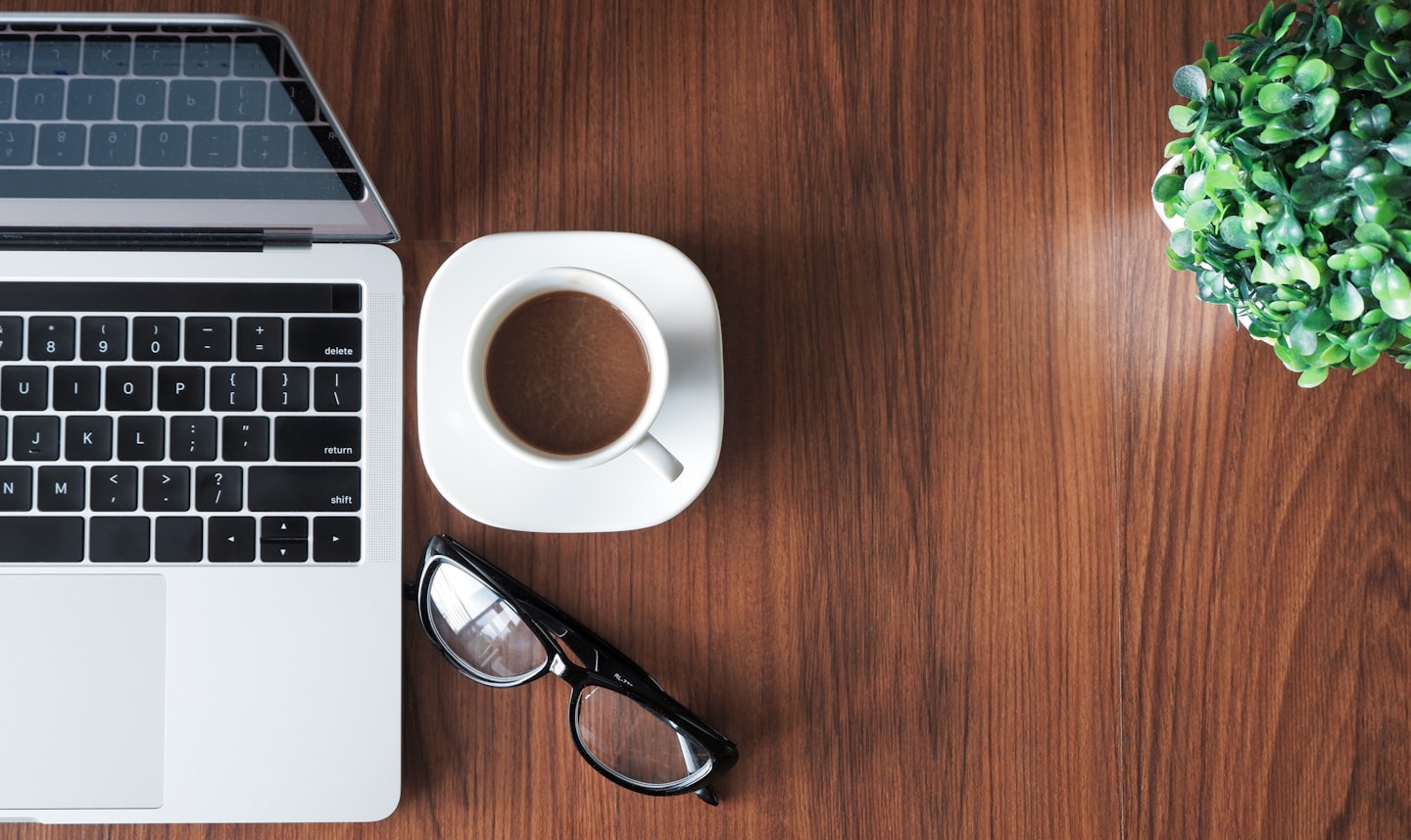Laptop, tazza di caffè, occhiali e pianta su una scrivania di legno.